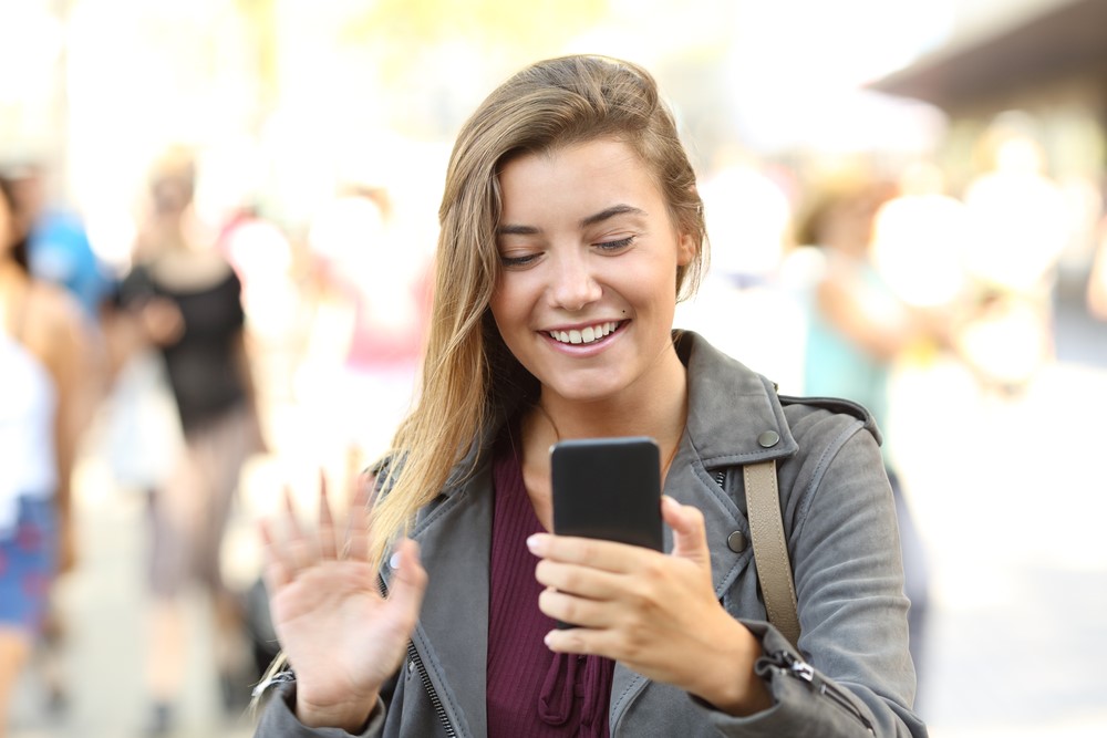 Frau grüßt auf der Straße freudig in ihr Smartphone